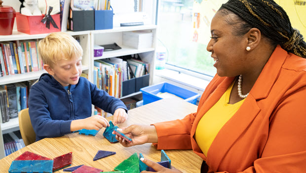 A teacher sits with a child at a table, guiding play with magnatiles.