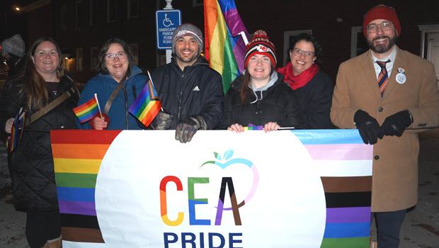 Teachers bundled up for the cold stand behind a CEA Pride banner outside at night.