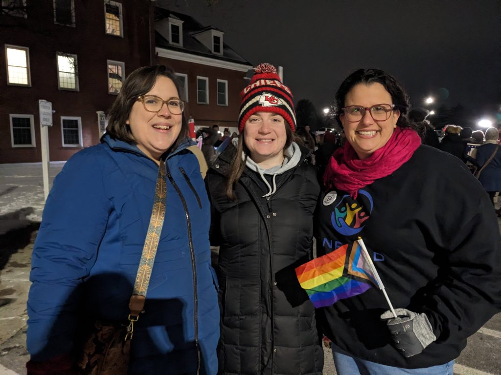Three smiling women stand outside at night. One holds a Pride flag. 